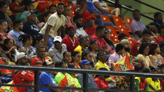 Fans in colorful attire and waving flags cheer during a Global Super League cricket match in a packed stadium.