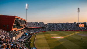 Wide-angle view of Emirates Old Trafford cricket stadium during a packed evening match, with spectators filling the stands under stadium lights.
