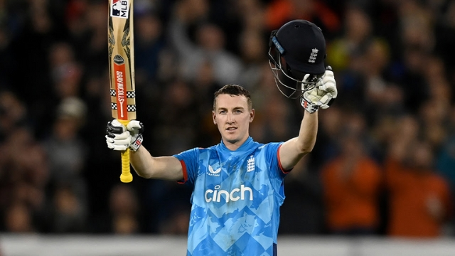 England cricket captain Harry Brook celebrates a century, raising his bat and helmet in front of a cheering crowd.
