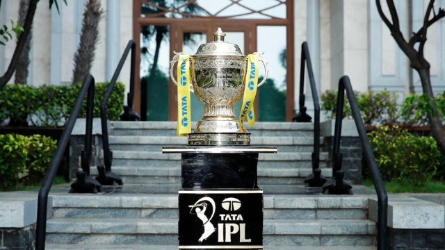 TATA IPL 2025 trophy displayed on a black pedestal outside a building, with yellow ribbons and official branding.