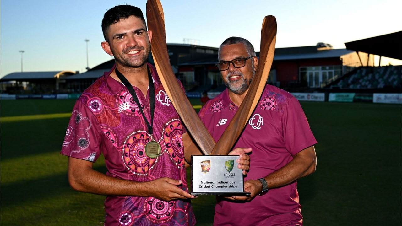 Jake Coolwell and Jason Smith holding the National Indigenous Cricket Championships trophy on the field, wearing maroon cultural jerseys.