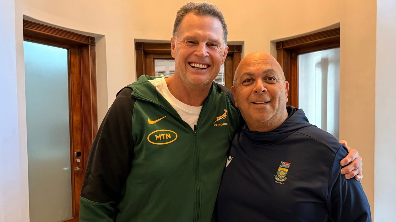 Rassie Erasmus and Shukri Conrad smiling together indoors, both wearing South African team apparel, representing rugby and cricket coaching leadership.