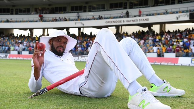 West Indies Test captain Roston Chase posing on the field with a red ball in hand after a match, in front of the Greenidge & Haynes Stand.