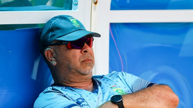 South Africa cricket head coach Shukri Conrad wearing team gear and sunglasses while seated in the dugout.