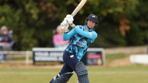 Ami Campbell playing a cricket shot in blue and navy team kit during a match, with focus on her batting stance and follow-through.