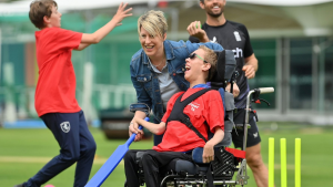 Child in a wheelchair joyfully playing cricket with assistance on a grass field, surrounded by smiling participants in a disability-inclusive cricket event.