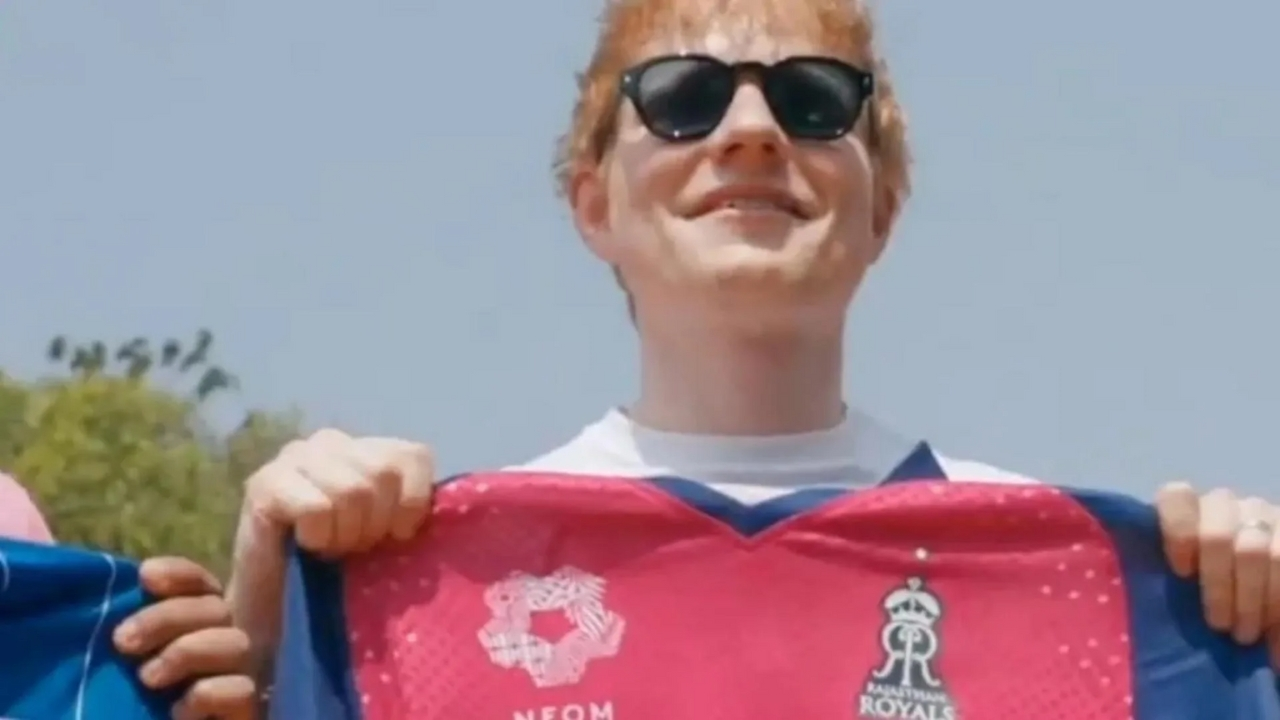 Ed Sheeran smiling while holding a Rajasthan Royals cricket jersey outdoors on a sunny day.