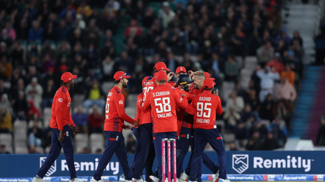 England men's cricket team players in red kits celebrating together on the field during a T20 match, with a crowd in the background.