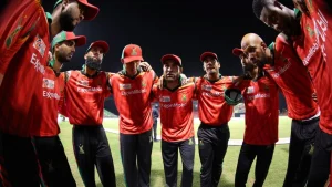 Guyana Amazon Warriors players in a team huddle during a Caribbean Premier League (CPL) 2024 match, wearing red jerseys under stadium lights.