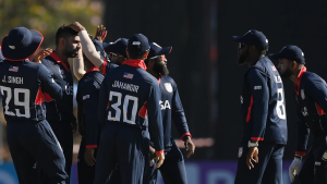 USA men's cricket team players celebrate together on the field during a match, wearing navy blue uniforms with player names and numbers visible.