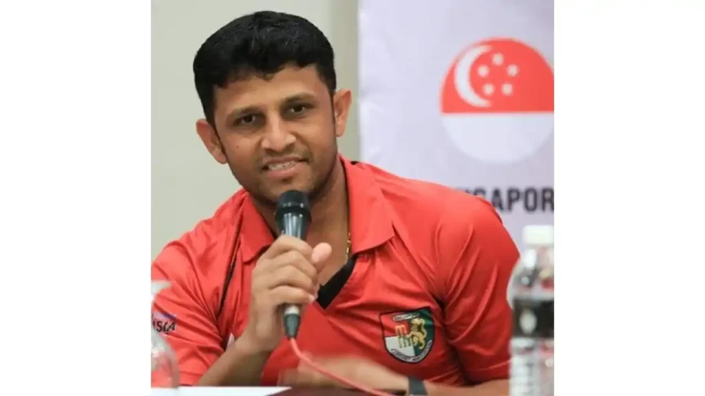 Chetan Suryawanshi speaking at a press conference, wearing a red Singapore cricket jersey, with a microphone in hand and a Singapore flag backdrop.