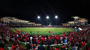 Packed crowd at a night CPL cricket match in Trinidad with fans dressed in red and stadium lights illuminating the field.