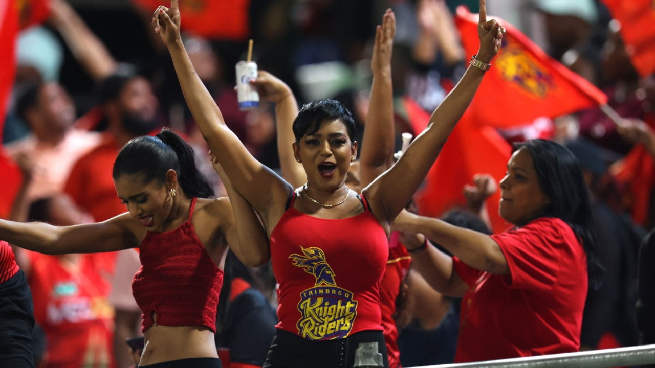 Trinbago Knight Riders fans enthusiastically cheer during a Caribbean Premier League (CPL) match, wearing team shirts and waving red flags.