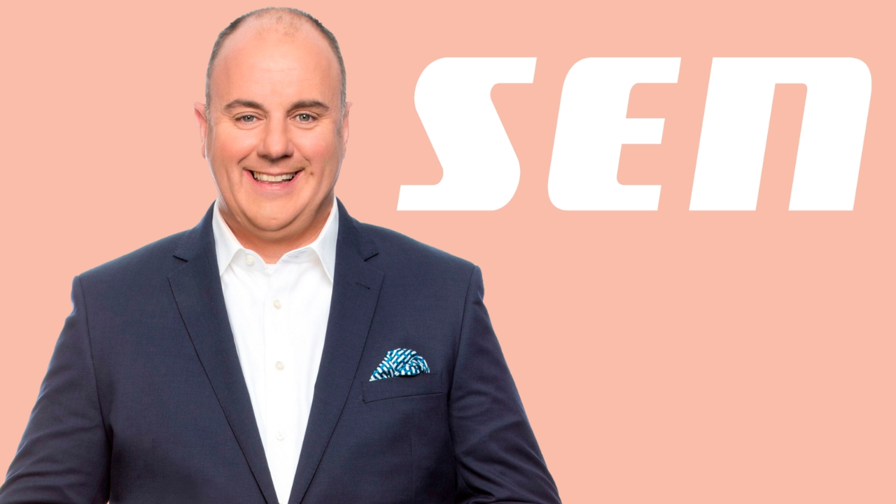 Craig Hutchison in a suit smiling in front of the SEN radio network logo on a peach background