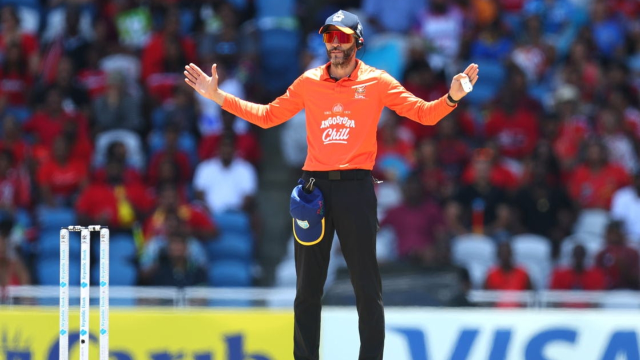 Cricket umpire in orange jersey signaling a wide ball during a Caribbean Premier League match, with crowd in the background.