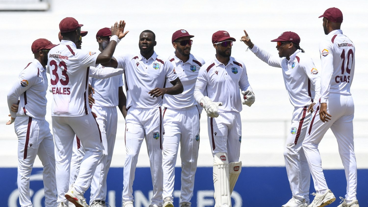 Cricket West Indies men's team wearing white Test jerseys celebrating a wicket on the field during a match, with players gathered in a huddle.