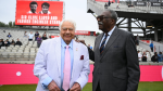 Farokh Engineer and Sir Clive Lloyd smiling together in front of the newly renamed "Sir Clive Lloyd and Farokh Engineer Stand" at a cricket stadium.