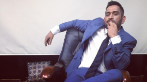 MS Dhoni sitting confidently in a chair wearing a blue suit and tie, exuding charisma and style.