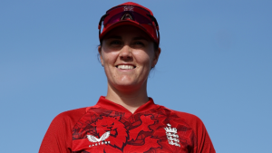 Nat Sciver-Brunt smiling in England women's red cricket jersey and cap, standing under a clear blue sky.