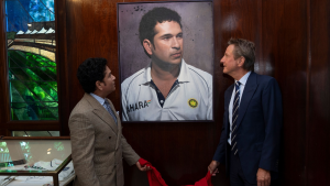 Sachin Tendulkar and Mark Nicholas unveiling a portrait of Tendulkar wearing an India cricket jersey, displayed on a wooden wall.