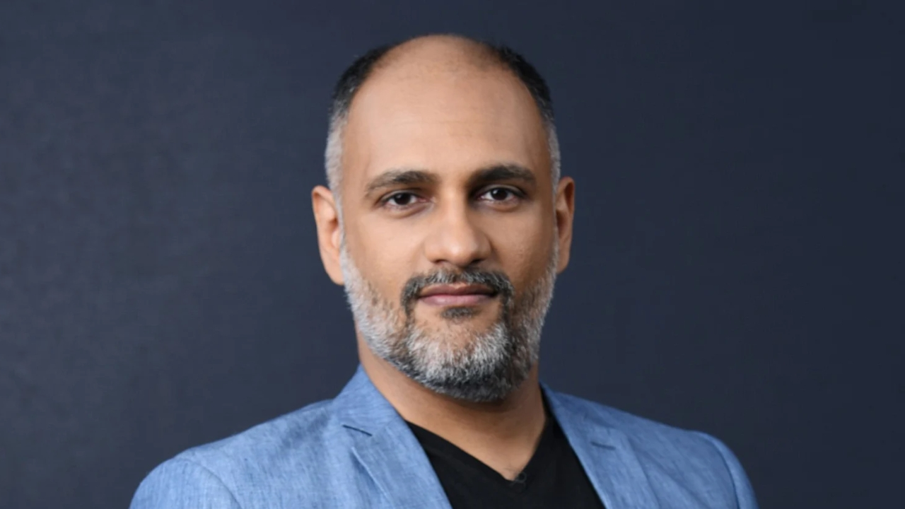Portrait of Sanjog Gupta wearing a light blue blazer and black shirt against a dark blue background.