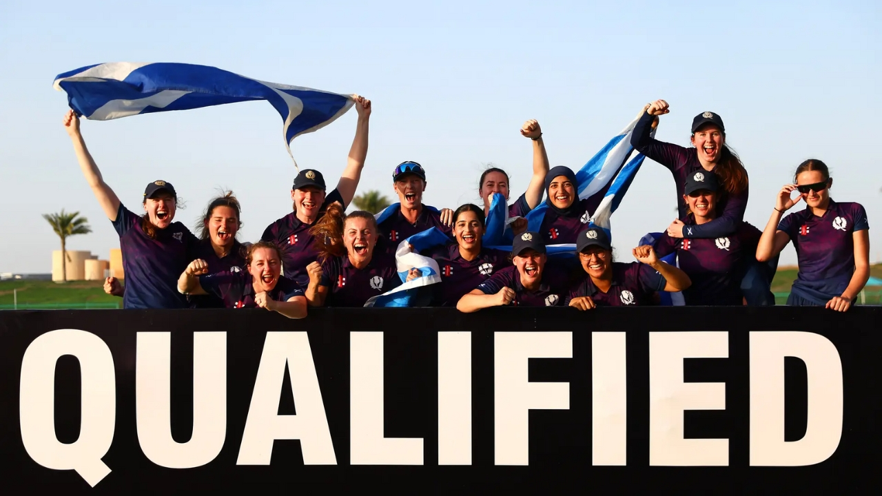 Scotland women's cricket team celebrates qualification with flags and cheers behind a large "QUALIFIED" sign.