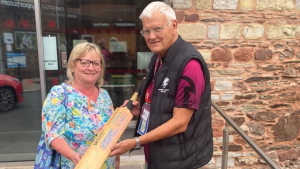 Man and woman holding a Sir Viv Richards cricket bat donated to the Somerset Cricket Museum outside a stone building.