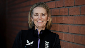 Charlotte Edwards, head coach of the England women’s cricket team, smiling while standing against a brick wall in official team attire.