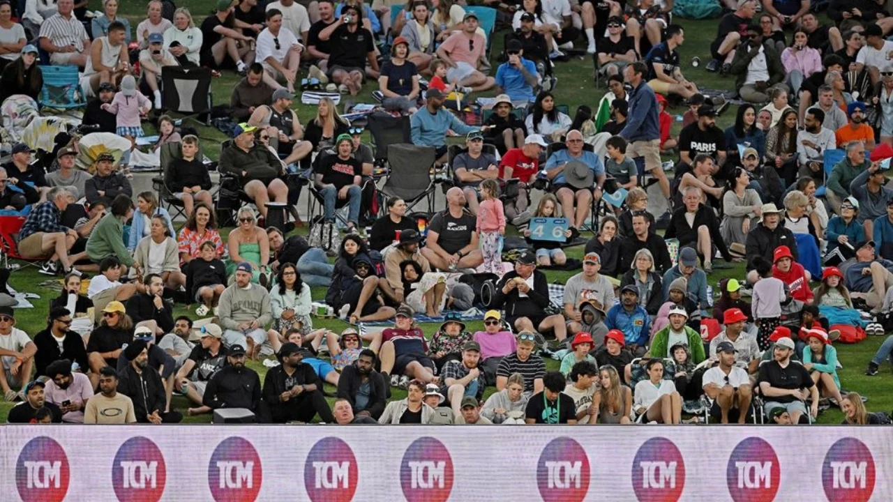 A large crowd of diverse cricket fans sitting on grass and chairs at a stadium during a match, enjoying the event in a relaxed atmosphere.