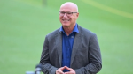 David White smiling while standing on a cricket field, wearing a grey blazer and blue shirt.
