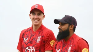 Harry Brook and Adil Rashid in red England cricket jerseys during a team event or practice session, smiling and standing side by side.