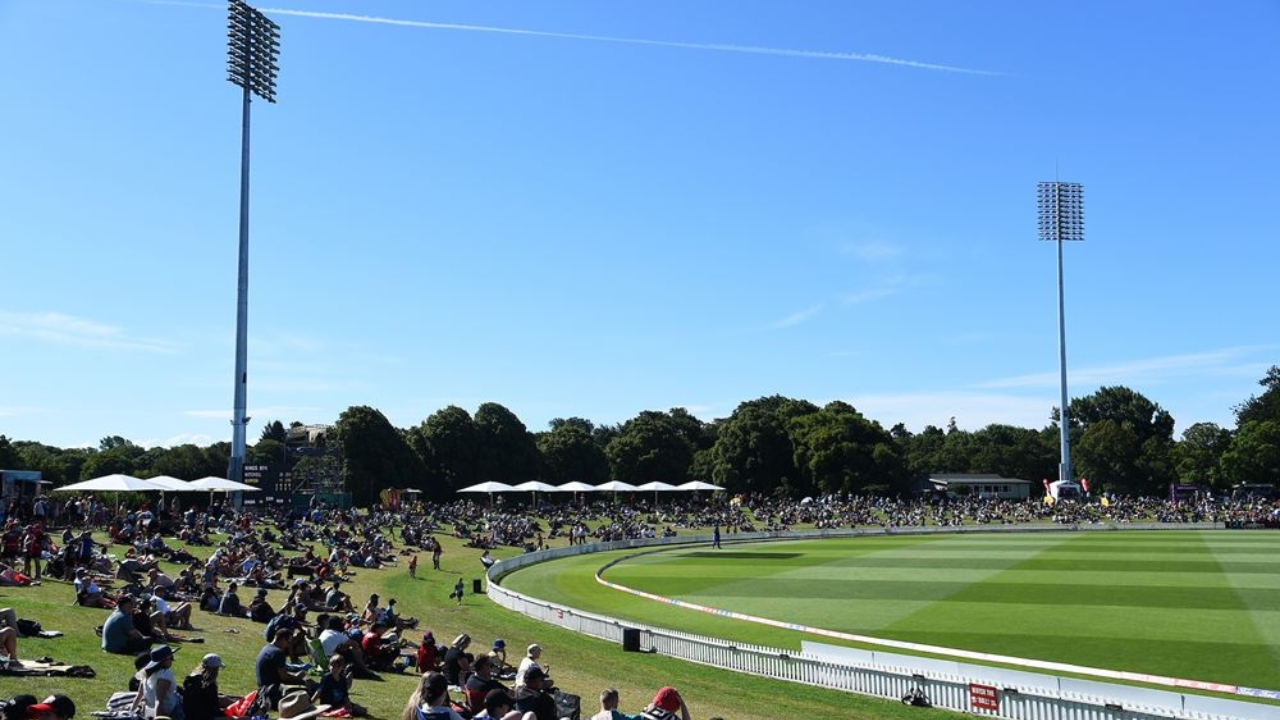 Spectators sitting on the grassy banks of Hagley Oval in Christchurch, New Zealand, watching a live cricket match under clear blue skies.