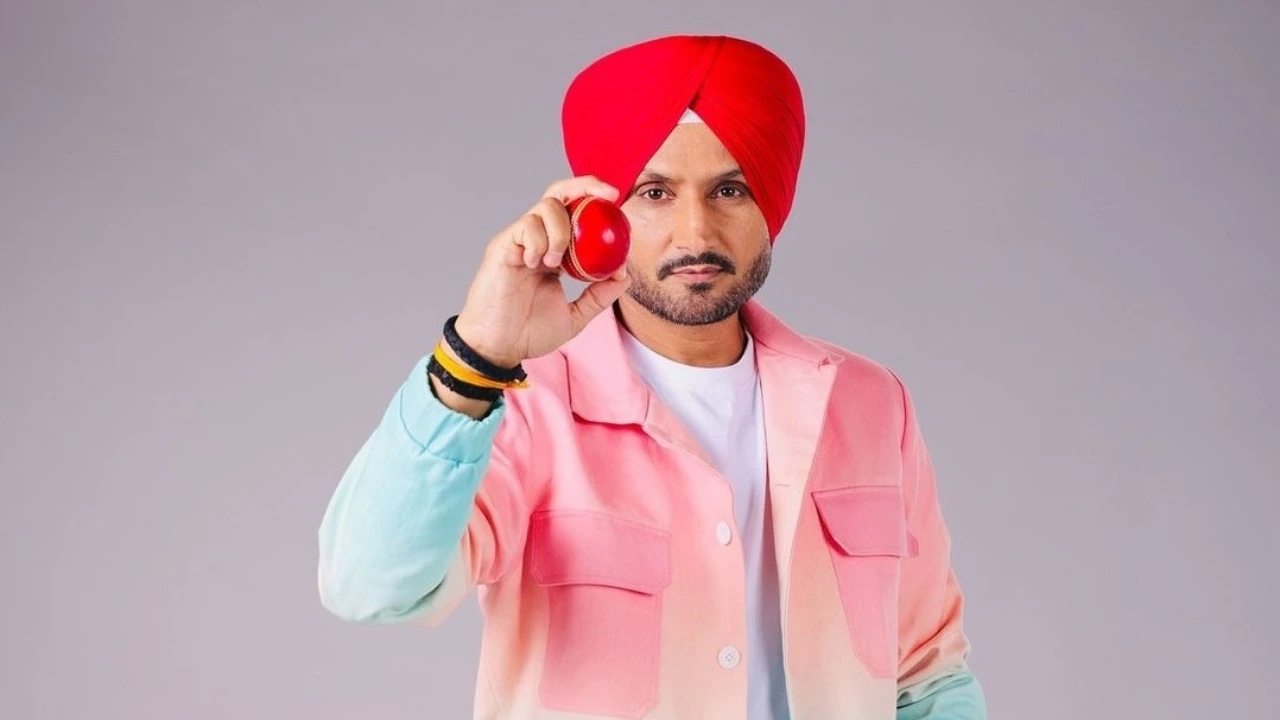 Harbhajan Singh wearing a red turban and colorful jacket holding a red cricket ball against a plain background.