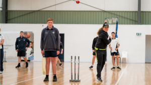 Students participating in an indoor cricket training session, with one student preparing to bowl and another holding a cricket bat in front of the stumps.