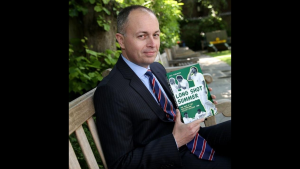 Neil Robinson, Head of Heritage and Collections at MCC, holding the book "Long Shot Summer" while seated on a wooden bench outdoors.