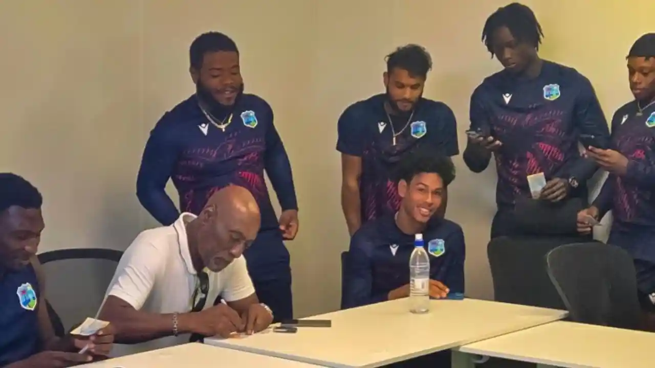 Sir Vivian Richards signing autographs while surrounded by young West Indies cricketers in team training gear.