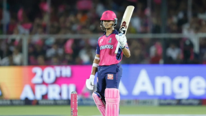 Yashasvi Jaiswal in Rajasthan Royals jersey holding his bat during an IPL 2025 match, with a colorful crowd and digital advertisement board in the background.