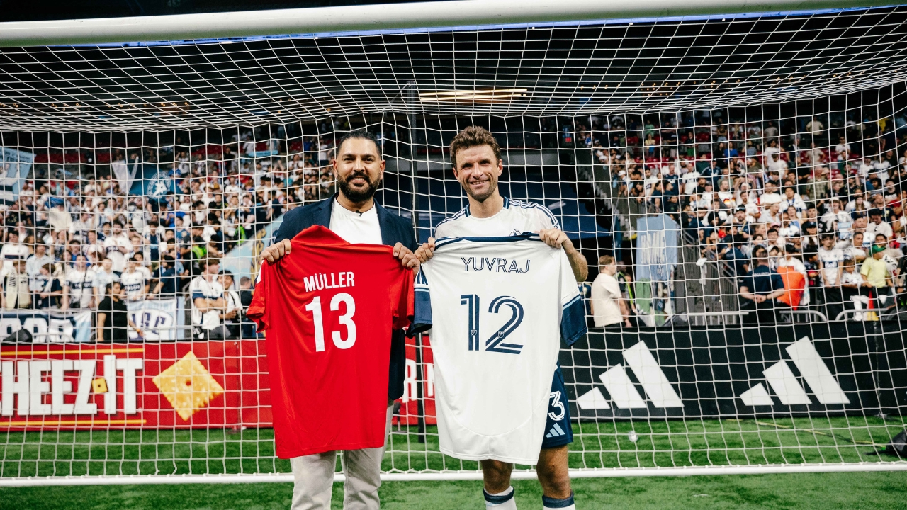 Yuvraj Singh and Thomas Müller exchange jerseys on a football field, holding shirts with each other's names and numbers in front of a goalpost and cheering crowd