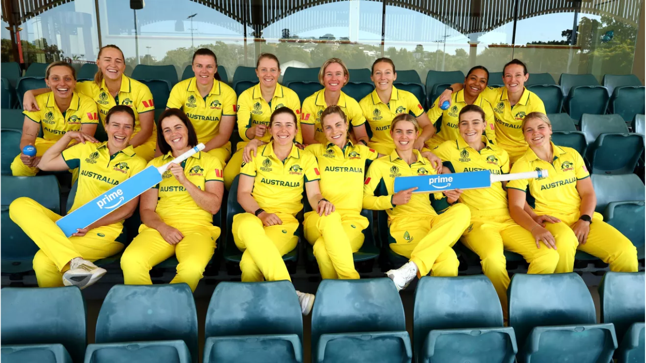 Australian women's cricket team sitting in stadium seats, smiling and holding a cricket bat with Amazon Prime branding.