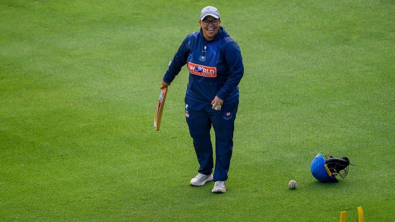 Claire Terblanche smiles during a cricket training session on a grass field, holding a bat with a helmet and ball on the ground nearby.