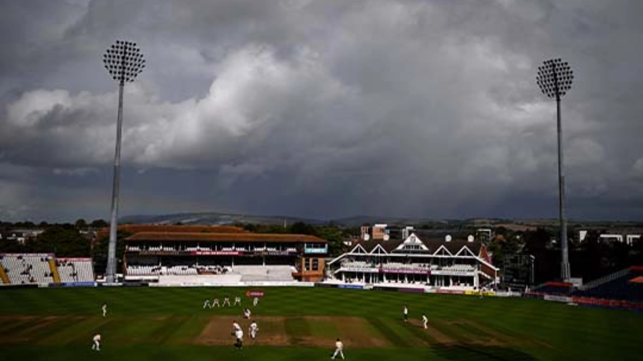 Aerial view of a cricket match in progress at a stadium under dark, stormy clouds with empty stands and floodlights