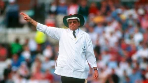 Legendary cricket umpire Dickie Bird signaling during a match, wearing a white coat and hat with a packed crowd in the background