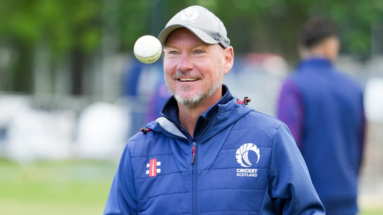 Doug Watson, head coach of Cricket Scotland, smiling while a cricket ball floats in the air during a practice session.