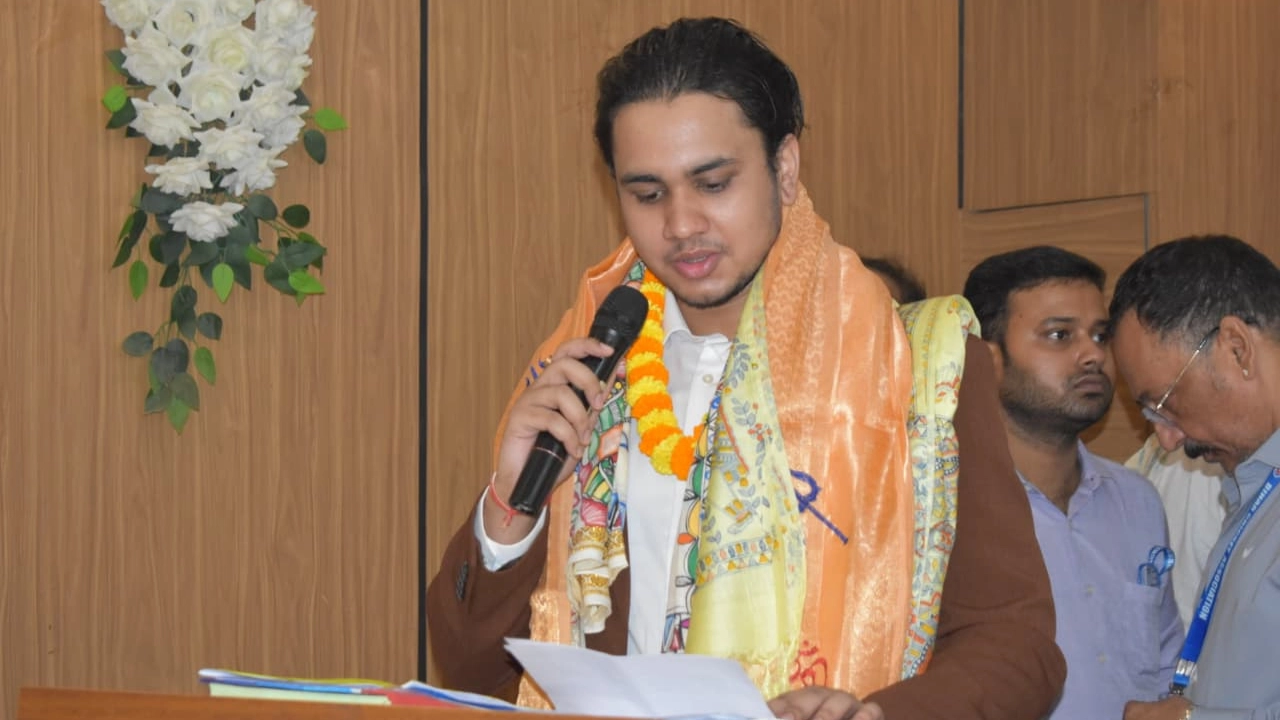 Harsh Vardhan speaking at an event, wearing a garland and traditional shawl, holding a microphone and reading a speech