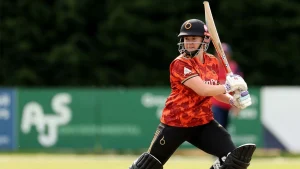 Kathryn Bryce playing a shot during a match in The Hundred 2025, wearing a red and orange team jersey with helmet and batting gear.