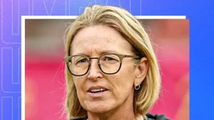 Close-up of Lisa Keightley, former Australian cricketer and coach, wearing glasses and standing in front of a blurred sports background