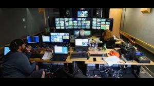 Technicians managing live sports feeds inside a broadcast control room with multiple screens and laptops.