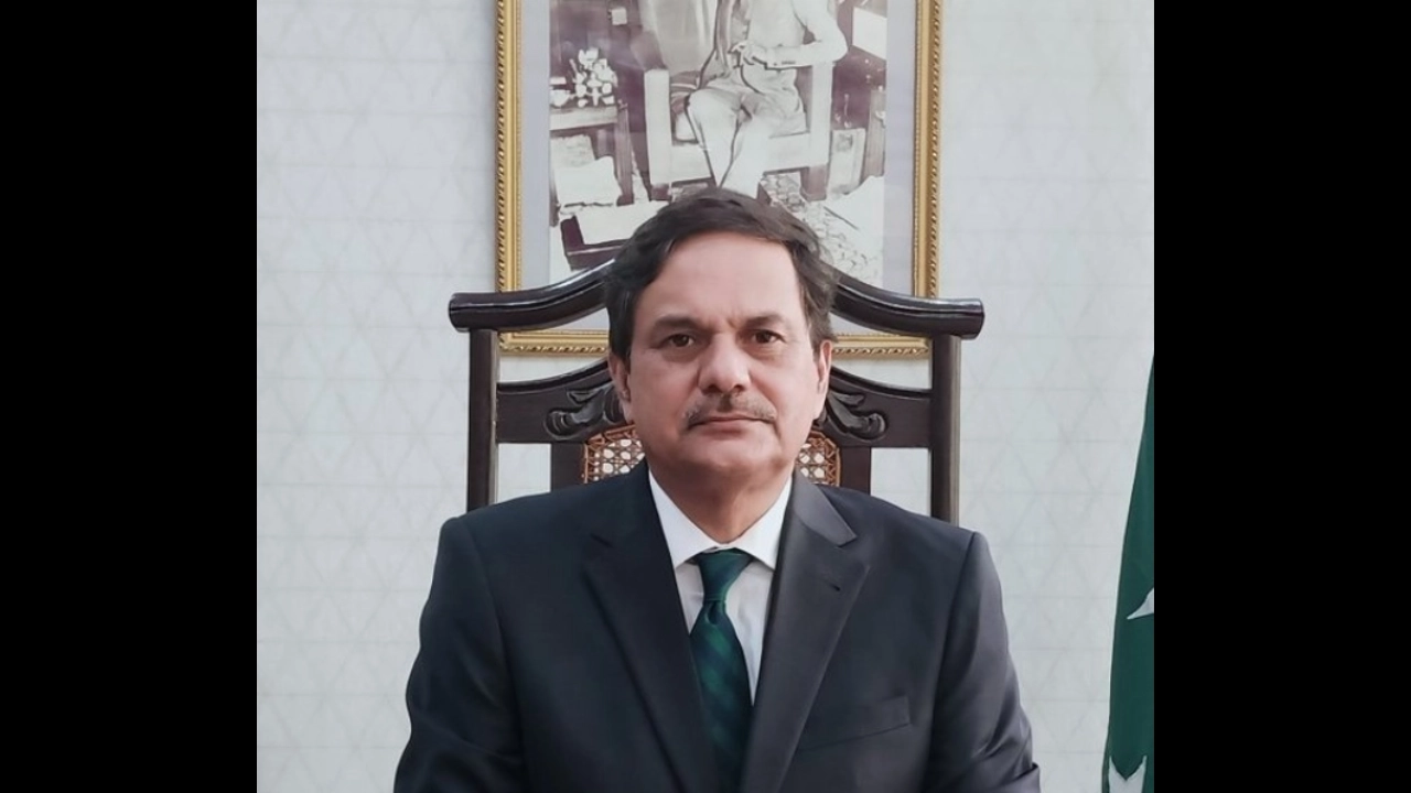 Portrait of Mansoor Qadir in formal attire seated in office with Pakistan flag and framed photograph in background