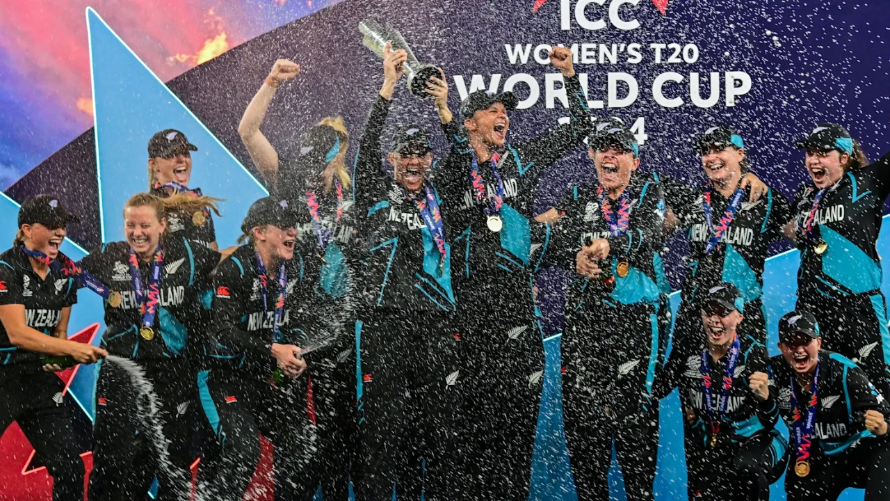 New Zealand women’s cricket team celebrating with champagne after winning the ICC Women’s T20 World Cup 2024, holding the trophy and wearing gold medals on stage.