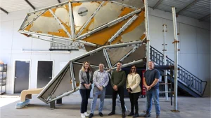 Group of five people standing in front of a large geometric metal sculpture inside an industrial workshop or fabrication facility